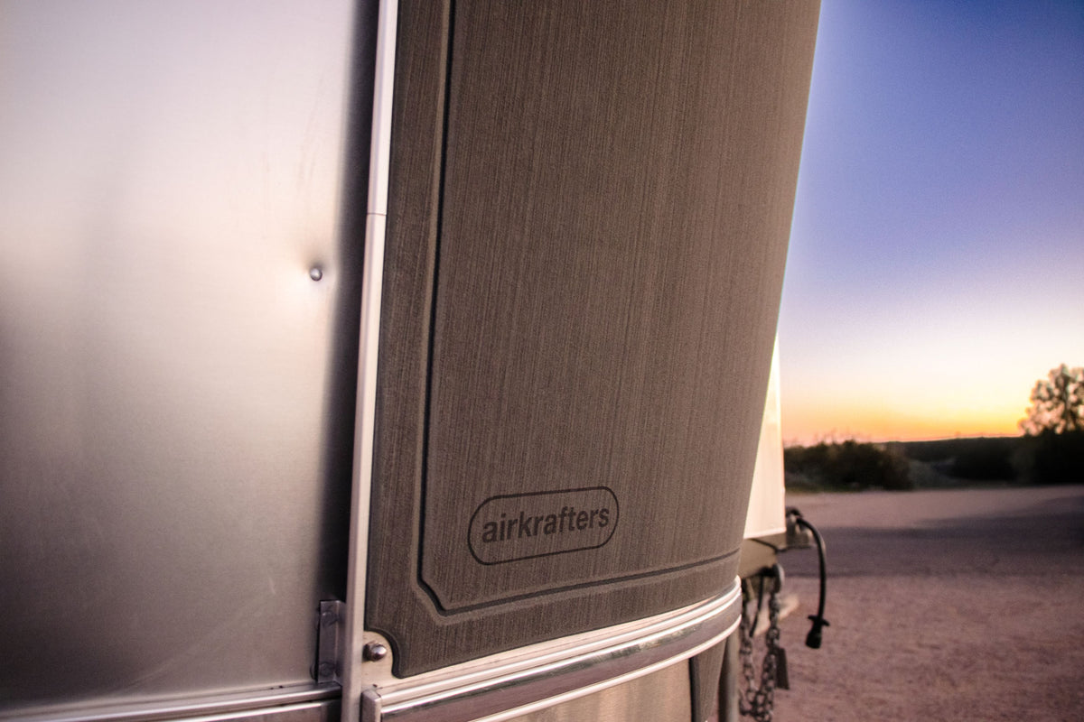 Close-up of an airstream trailer at sunset, showing the word airkrafters on its side, with a scenic background of desert and trees under a colorful sky.