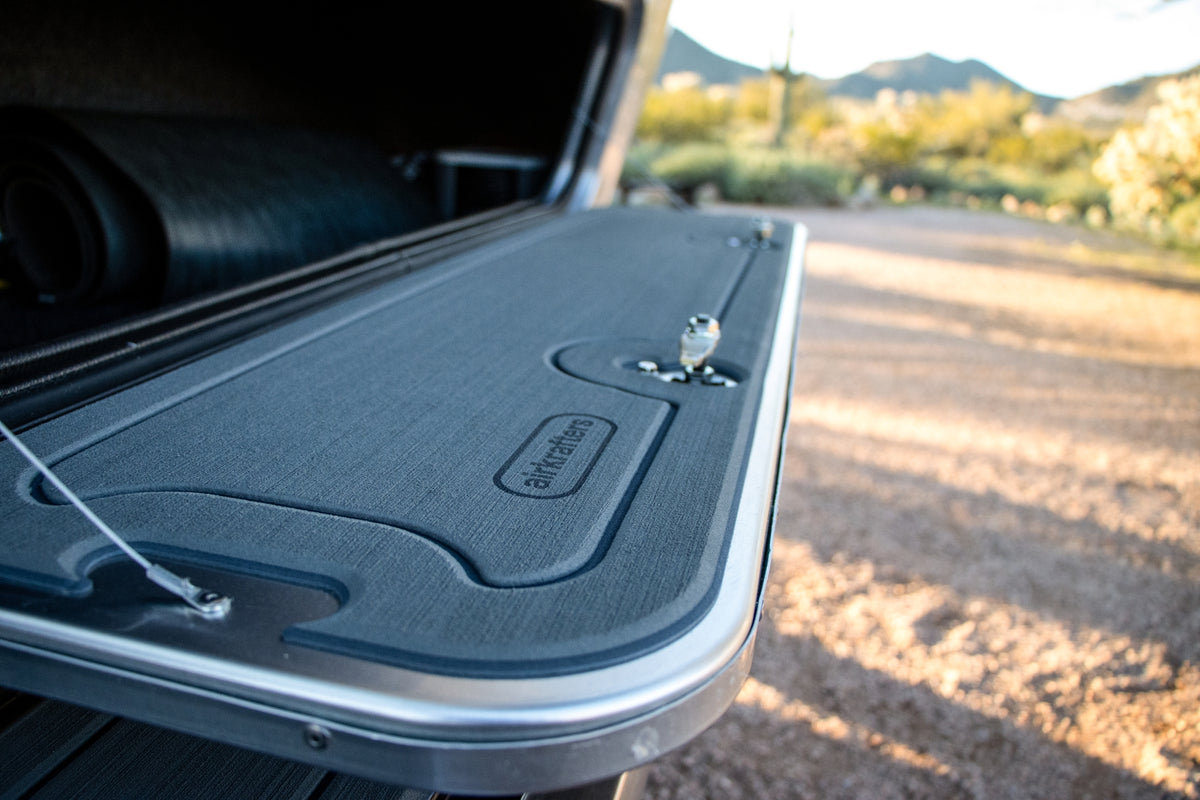 Close-up of a truck bed cover partially open in an outdoor setting, revealing a rolled-up dorMAT™ by Airkrafters and an EVA foam liner inside, with a desert landscape in the background.