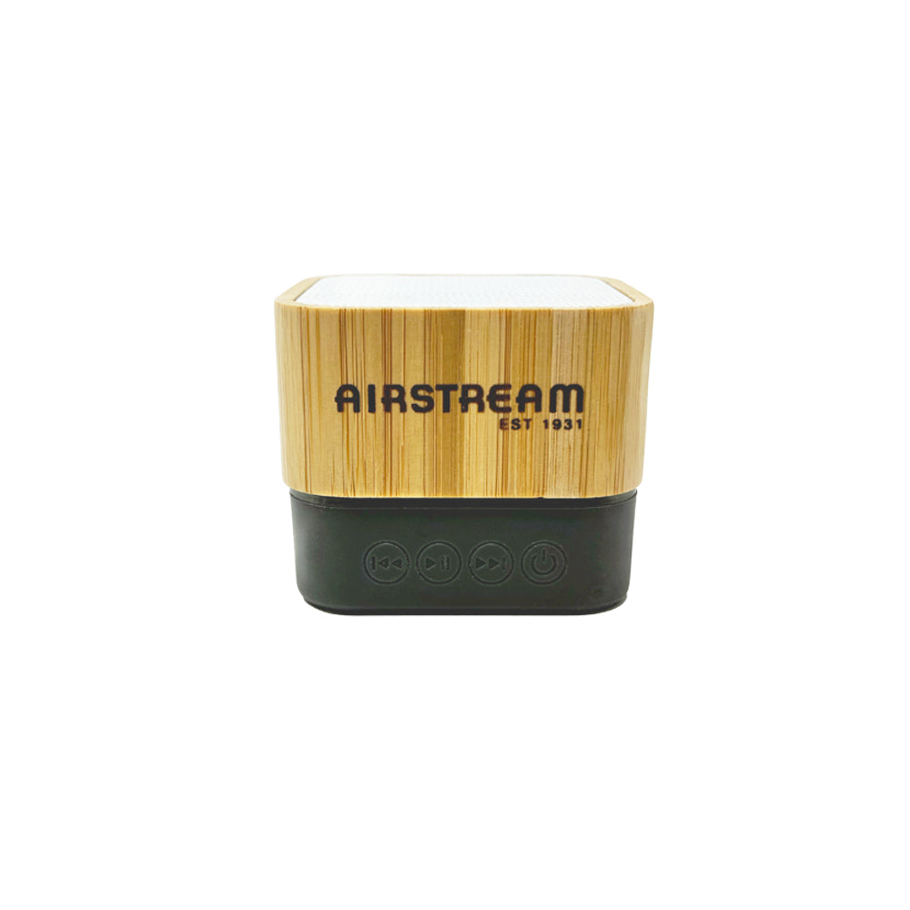 A compact, square Airstream 1931 Art Deco Bamboo Wireless Light Up Speaker features a bamboo top and a black base, with “AIRSTREAM” displayed on the front. This eco-friendly portable speaker offers control buttons—play, pause, and power—on the base.