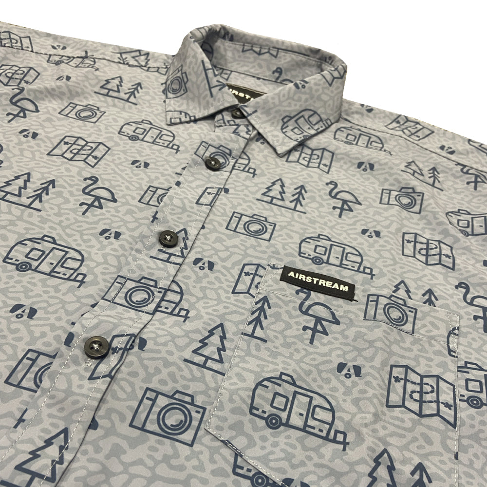 The Airstream Conversational Button Down Shirt is a lightweight gray shirt adorned with blue outlined patterns of cameras, trees, camper vans, and tents—making it the perfect choice for your next summer adventure. A black AIRSTREAM tag sits just above the chest pocket.