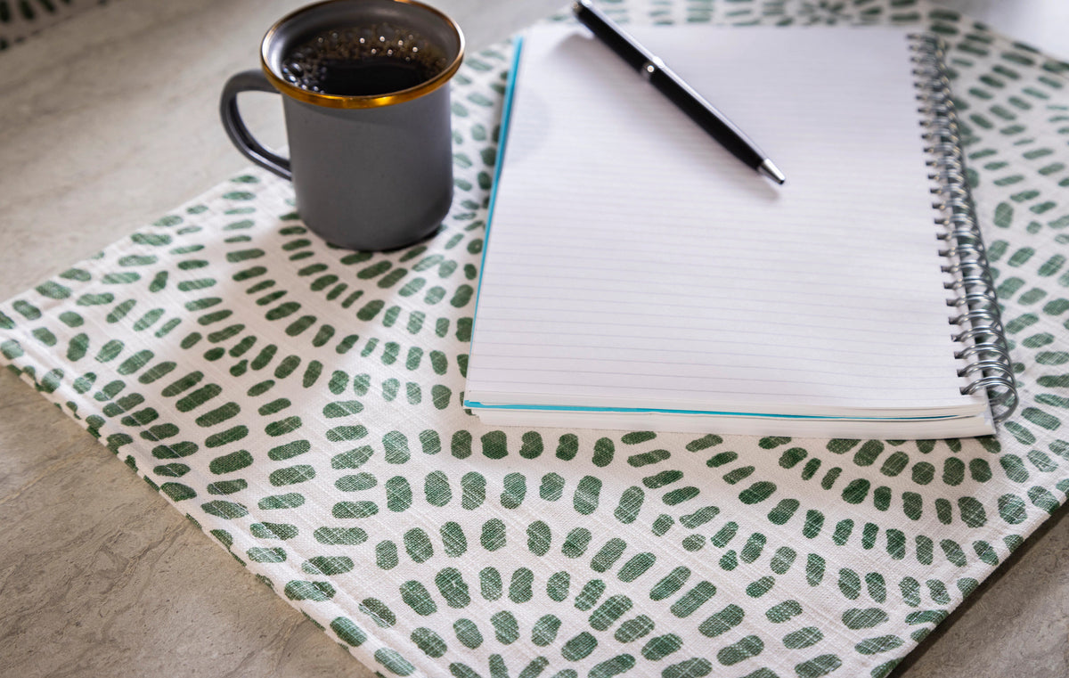 A gray mug filled with a dark beverage sits next to a spiral notebook and pen on a white mat with a green pattern, perfectly complementing the Placemat Set of 2 Bundle for a cohesive tabletop look.