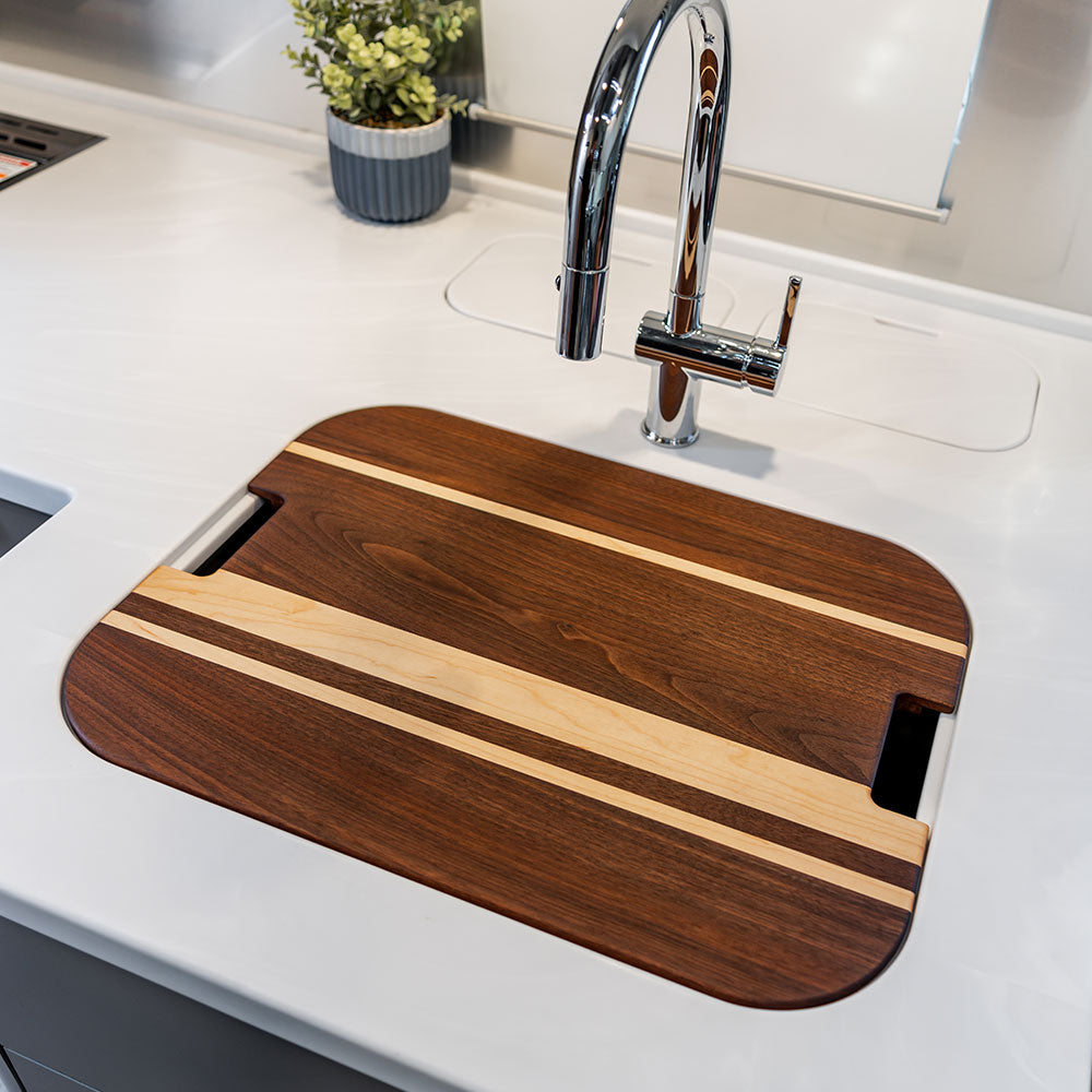 An Airstream Custom Sink Cutting Board for Classic Travel Trailers, featuring light and dark stripes, covers a sink beneath a modern chrome faucet in the kitchen, with a small potted plant visible in the background.
