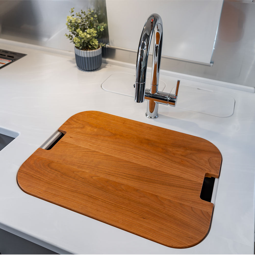 An Airstream Custom Sink Cutting Board for Classic Travel Trailers rests atop a white kitchen sink beneath a chrome faucet, combining modern style and practical functionality. In the background, a small potted plant adds a fresh touch of greenery—creating an ideal space for Airstream owners seeking accessories that integrate seamlessly with contemporary kitchen decor.