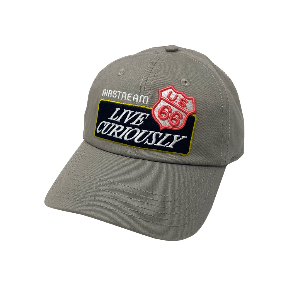The Airstream Route 66 Live Curiously Hat is a gray baseball cap adorned with embroidered patches featuring “AIRSTREAM,” a red U.S. Route 66 shield, and “LIVE CURIOUSLY” on a navy background. Designed with a curved brim and prominent stitching, this road trip hat is perfect for showcasing your adventurous spirit wherever your journey leads.