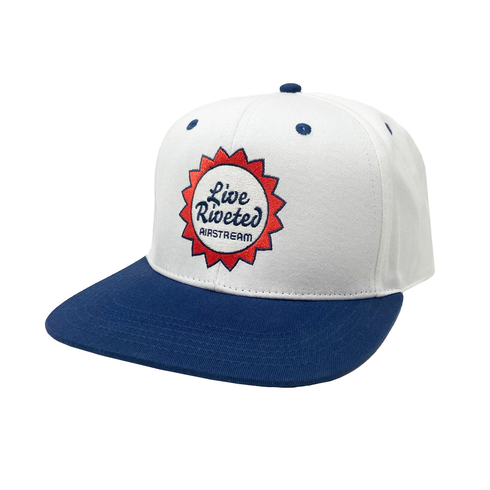 The Airstream Live Riveted Sun Trucker Hat features a white and blue design with a round red and blue embroidered patch on the front that reads “Live Riveted Airstream”—the perfect adventure hat for sunny days.