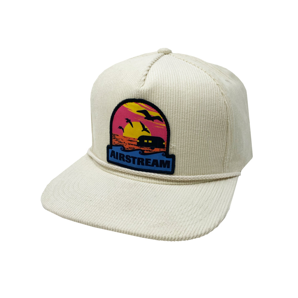 Introducing the Airstream Sunset Corduroy Trucker Hat—a cream corduroy hat designed with a vibrant patch showcasing an Airstream trailer, palm trees, and birds against a sunset backdrop. The patch features “AIRSTREAM” printed in blue, making this the ultimate road trip accessory for adventure seekers.