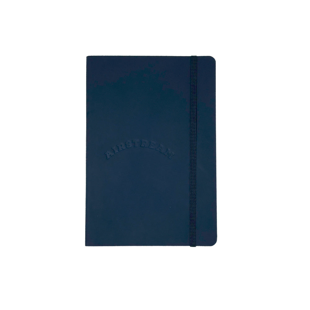 A closed Airstream Heritage Arc Soft Cover Journal in dark blue, featuring a soft cover with rounded corners and an elastic band closure, is displayed against a plain white background. The word “AIRSTREAM” is embossed on the cover, and the portable notebook contains lined pages inside.