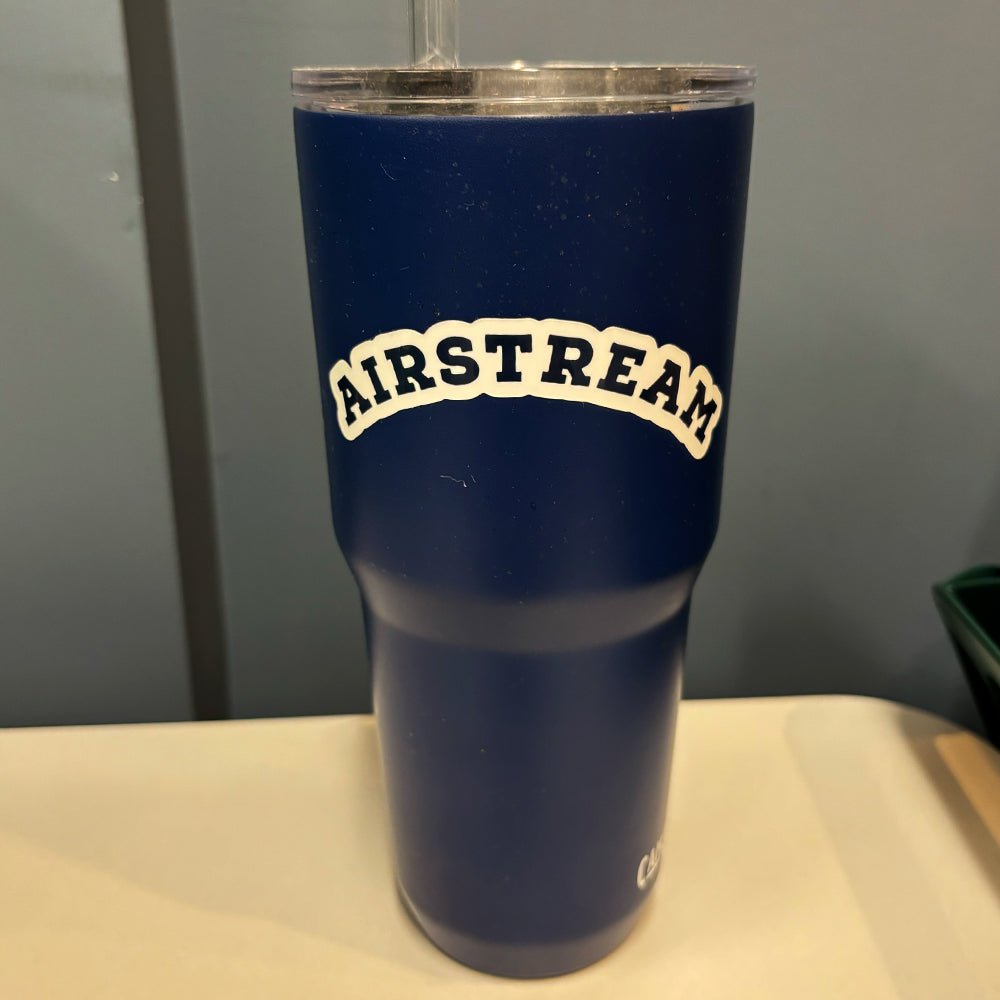 A blue tumbler with a clear lid and straw sits on a table. Featuring the Airstream Heritage Arc Sticker in bold, arched white letters on the front, it adds a touch of classic vintage style—a stylish addition to any travel gear collection.