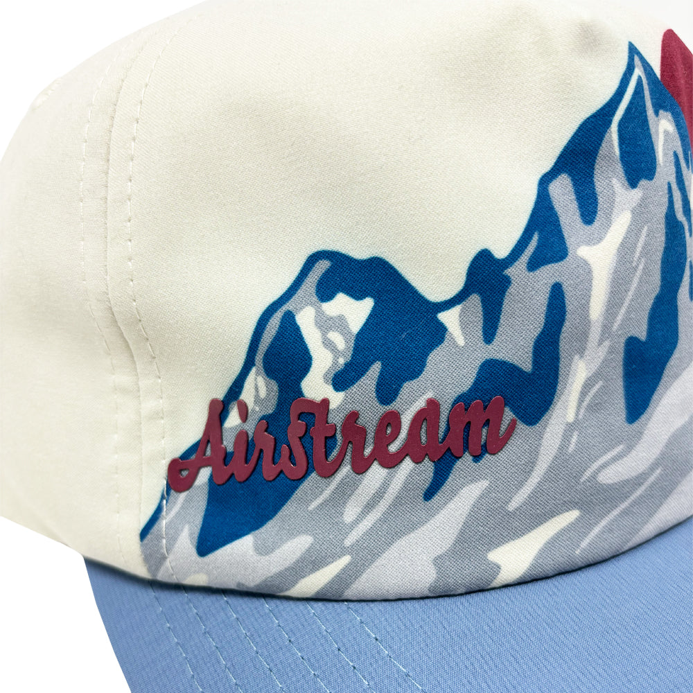 A close-up of the Airstream Mountain Peak Trucker Hat, featuring a white and blue design with a mountain graphic and “Airstream” embroidered in red script on the front.