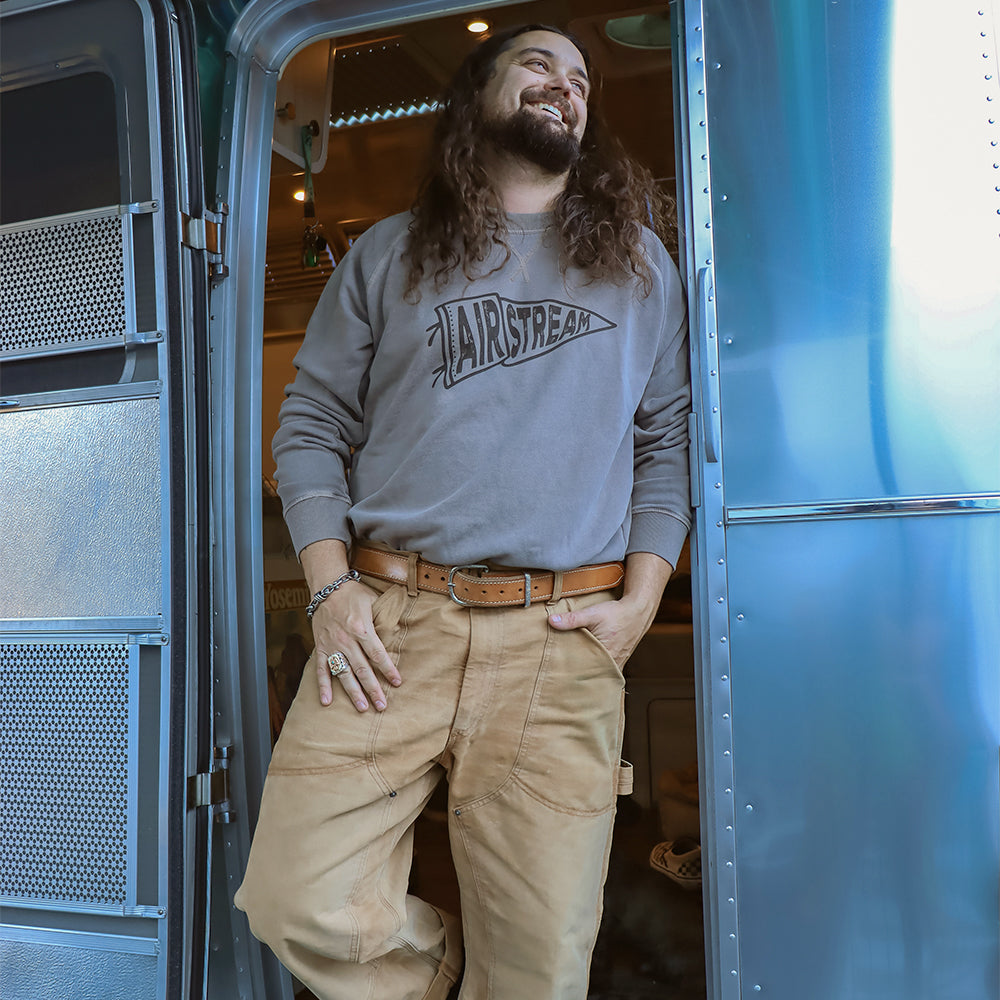 A man with long hair and a beard smiles as he stands in the doorway of a shiny blue trailer, wearing tan pants and the Airstream Rally Pennant Sweatshirt—ready for campfire gatherings under the stars.