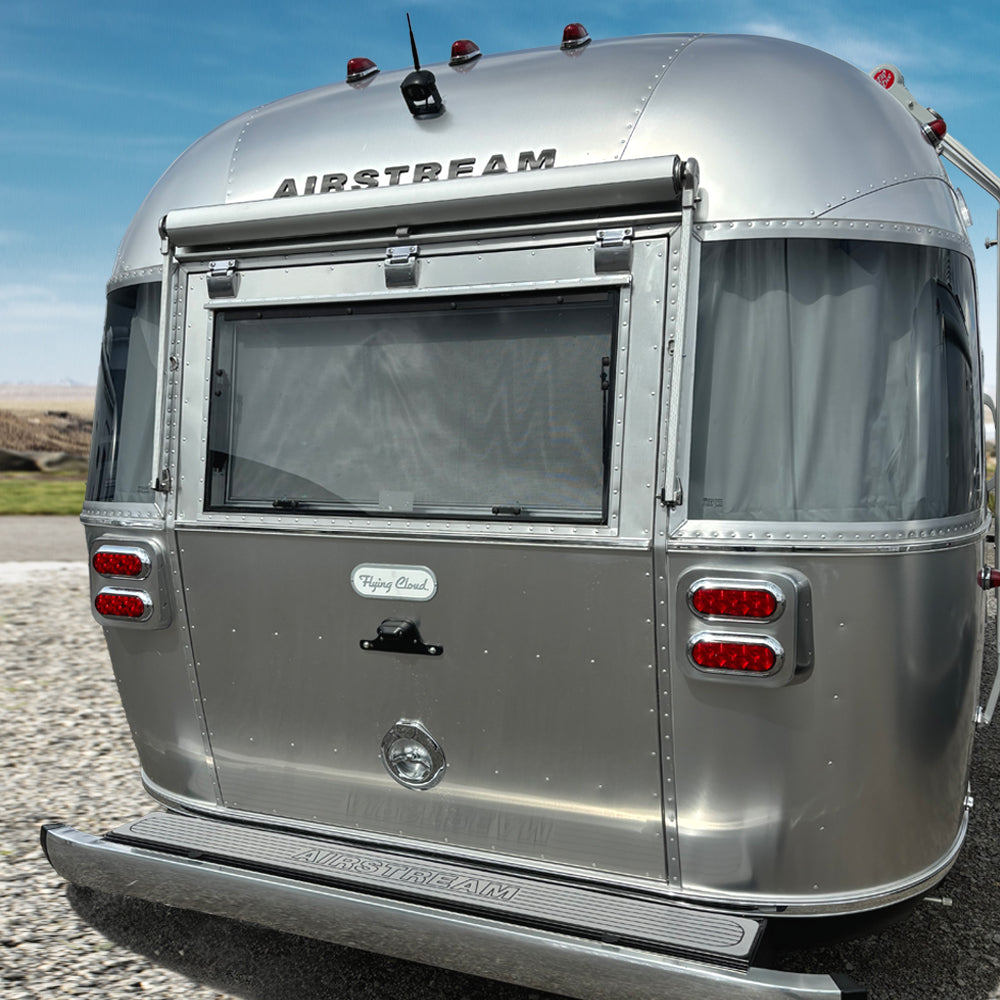 A silver Airstream travel trailer is parked on gravel with its rear window open, revealing red tail lights, a license plate area, an Airstream rear bumper fitted with the Airstream Rear Bumper Non-Slip Cover (Part #703938), and the Flying Cloud logo. The sky and fields are visible in the background.