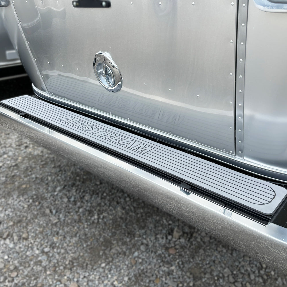 A close-up of an Airstream trailer’s silver door and step featuring the Airstream Rear Bumper Non-Slip Cover (Part #703938) installed, with “AIRSTREAM” printed on the cover. The step sits above small gravel stones, highlighting the enhanced safety and functionality of this accessory at the entrance.