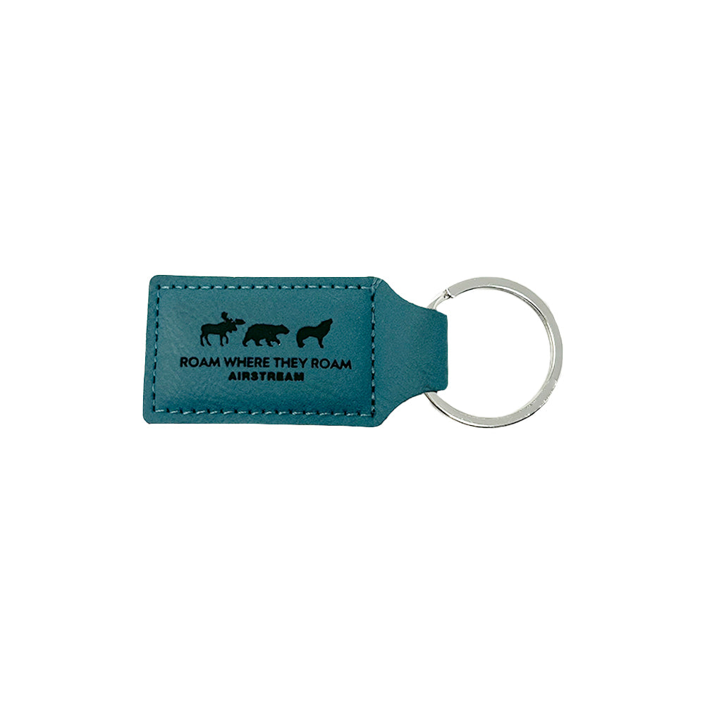 The Airstream Roam Where They Roam Keychain is crafted from blue leather and features silhouettes of a bison, bear, and wolf above the phrase “ROAM WHERE THEY ROAM AIRSTREAM.” It comes with a silver metal key ring attached on the right side and is an ideal gift for adventure-loving wanderers. Shown against a white background.