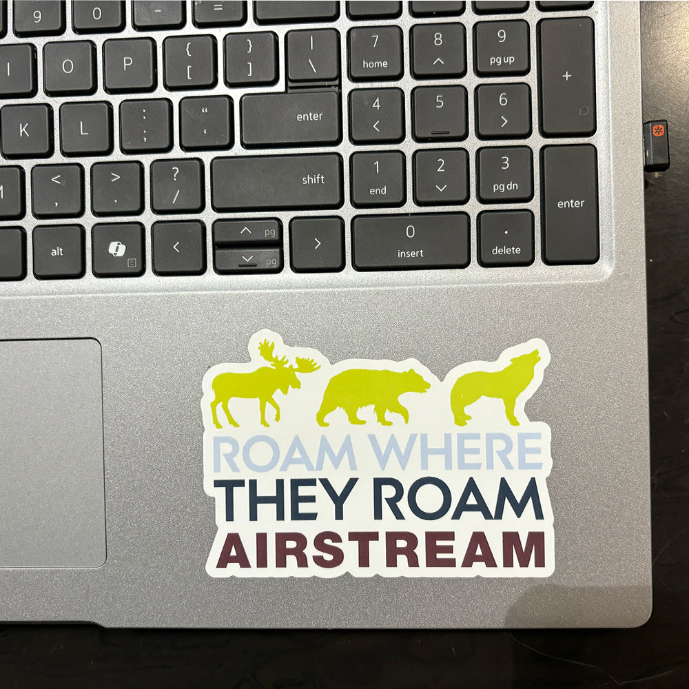 A laptop keyboard featuring the Airstream Roam Where They Roam Sticker, which displays silhouettes of a moose, bear, and wolf above the words “ROAM WHERE THEY ROAM AIRSTREAM.”.