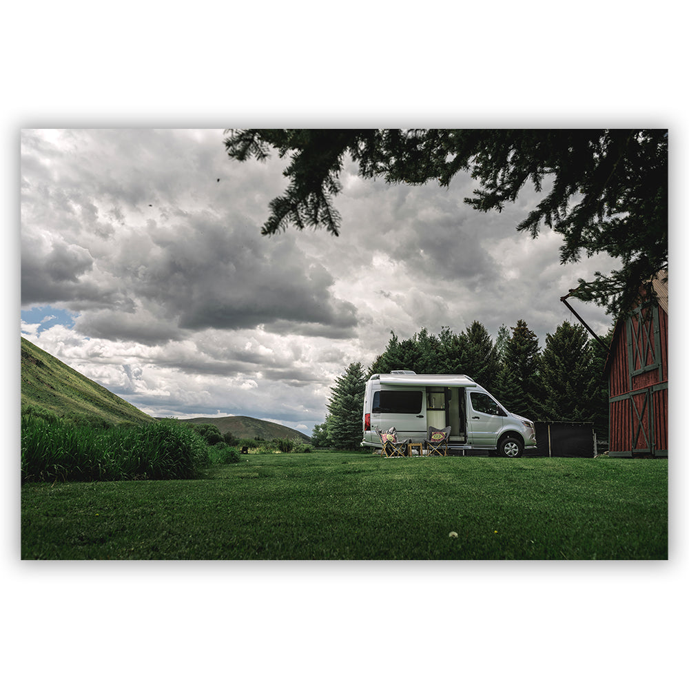 A white Airstream Touring Coach Picturesque Postcards is parked on green grass beside a red barn, with folding chairs arranged outside. Trees and hills surround the scene under a cloudy sky, evoking an image straight from a postcard.
