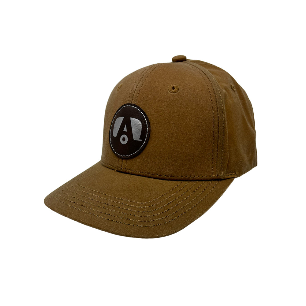 Airstream Trailer A Leather Patch Wax Cotton Hat features a vintage-style brown waxed cotton construction with a classic curved brim. This hat showcases a round black leather patch on the front, adorned with a stylized white A logo. Visible stitching, two ventilation eyelets, and a subtle distressed finish add unique character to this timeless accessory.