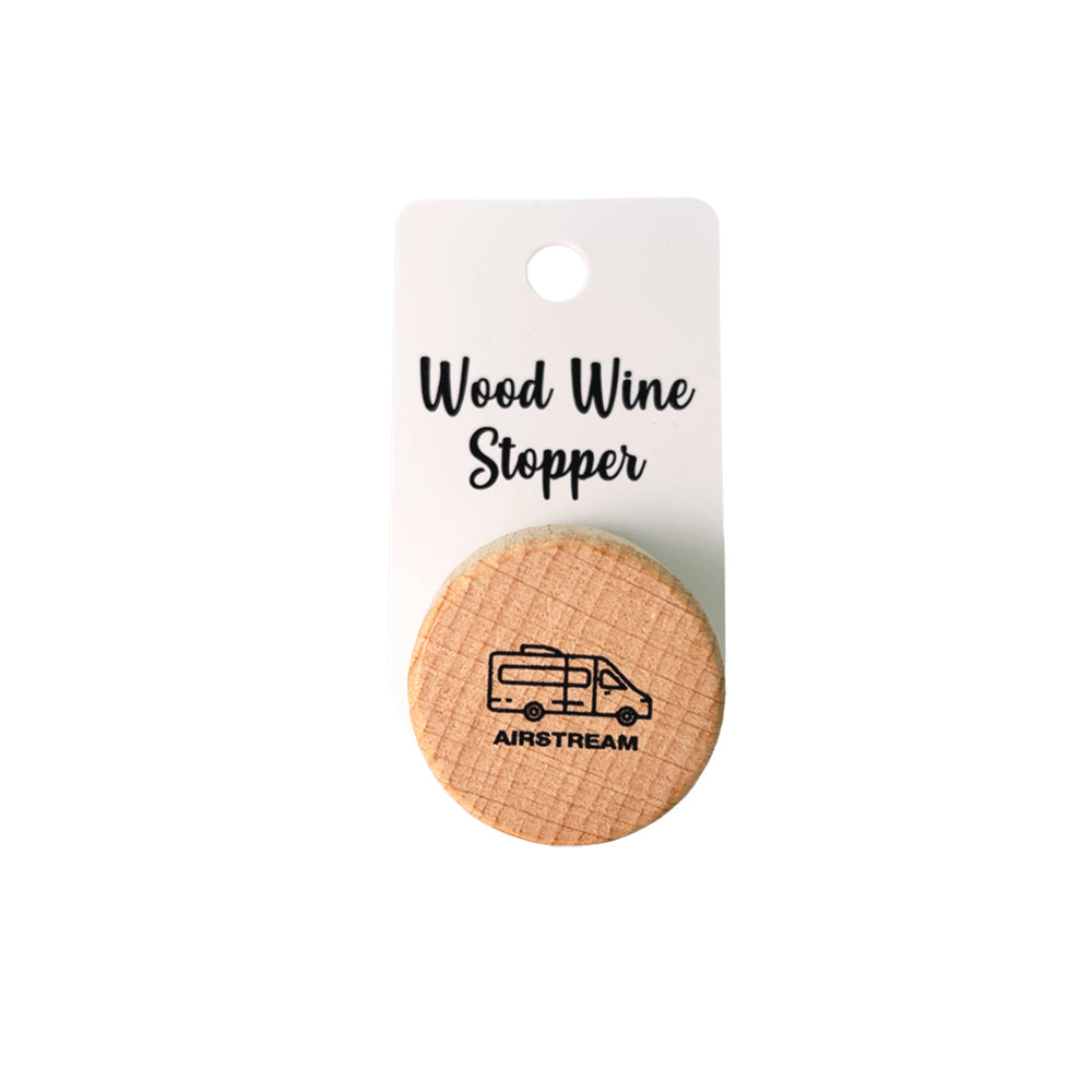 Introducing the Airstream Wine Stopper—a round wooden wine accessory featuring an illustration of a camper van and the word “AIRSTREAM,” attached to a card labeled “Wood Wine Stopper.”.
