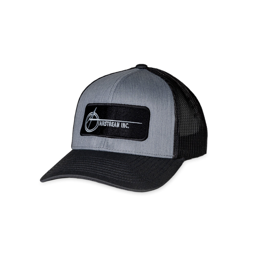The Airstream Space A Trucker Hat is a gray and black hat featuring a curved brim and mesh back. The front displays a rectangular patch with the official Airstream logo, showcasing the vintage Space A design and the text “AIRSTREAM INC.” in white.