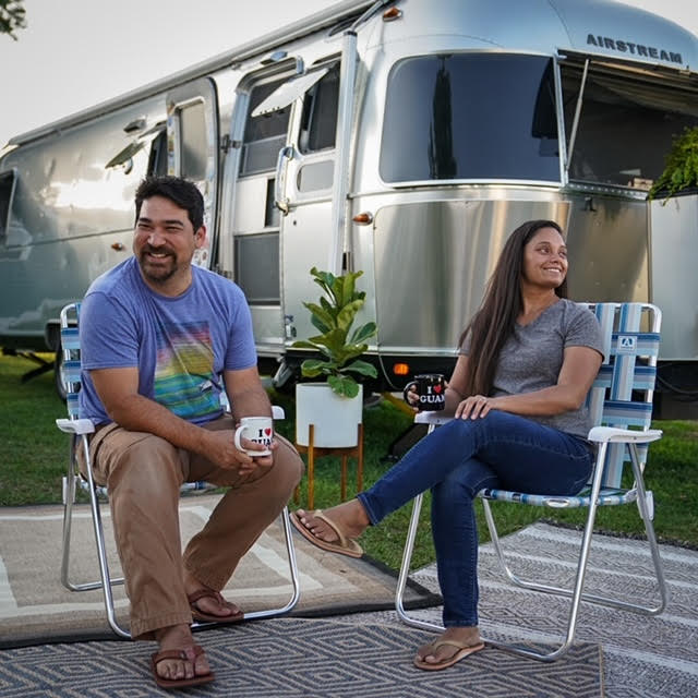 Two people sit outdoors on The Airstream Backtrack Chair by KUMA (Set of 2), smiling and holding mugs in front of a shiny silver Airstream trailer with a potted plant nearby.