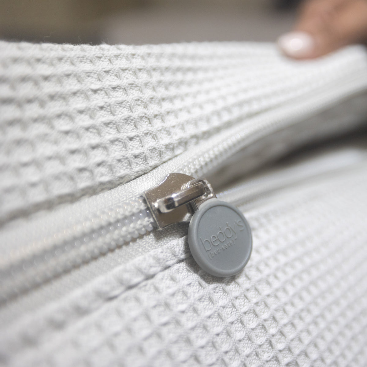 Close-up of a gray zipper on textured white fabric, featuring a round zipper pull labeled beddis. In the blurred background, a hand with painted nails adds an elegant detail—showcasing the thoughtful design of the Airstream Custom Fit Beddy’s for Quiksilver Travel Trailers bedding.
