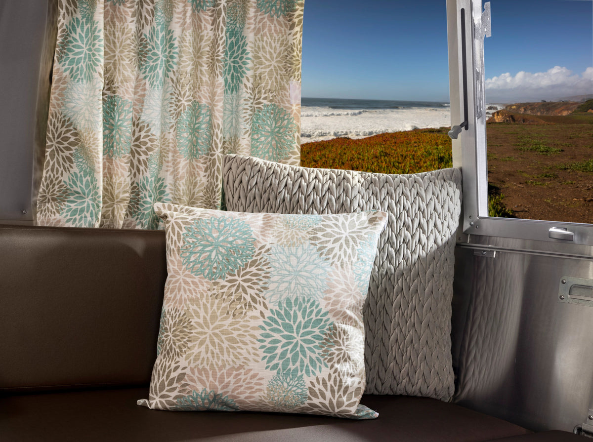 Throw pillows and a floral-patterned curtain sit on a bench in an RV, with an open window framing a coastal landscape of waves and blue sky—perfectly complemented by Airstream Printed Custom Curtains for Sport Travel Trailers.