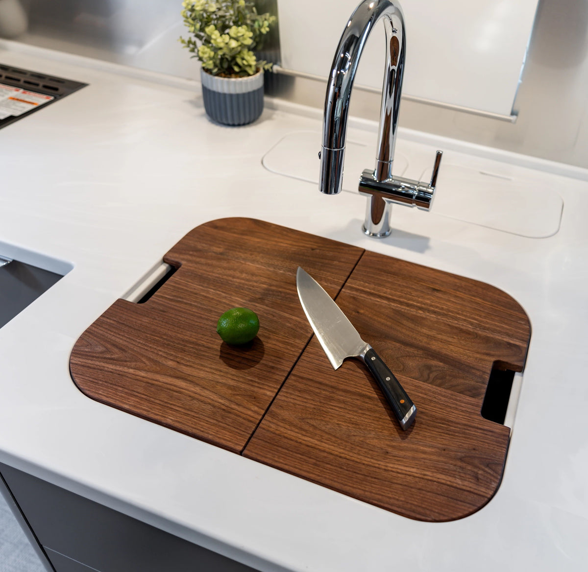 An Airstream Custom Sink Cutting Board for Pottery Barn Travel Trailers is placed over the sink, holding a chef’s knife and a lime. A chrome faucet and a small potted plant are visible on the white countertop.
