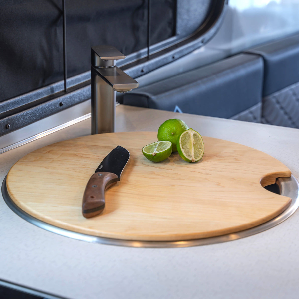 A wooden, round cutting board with a knife and two limes—one sliced in half—rests atop an Airstream Custom Sink Cutting Board for Basecamp in a sleek, modern kitchen, making it the perfect accessory for anyone wanting custom sink cutting boards or essential Airstream Basecamp accessories.