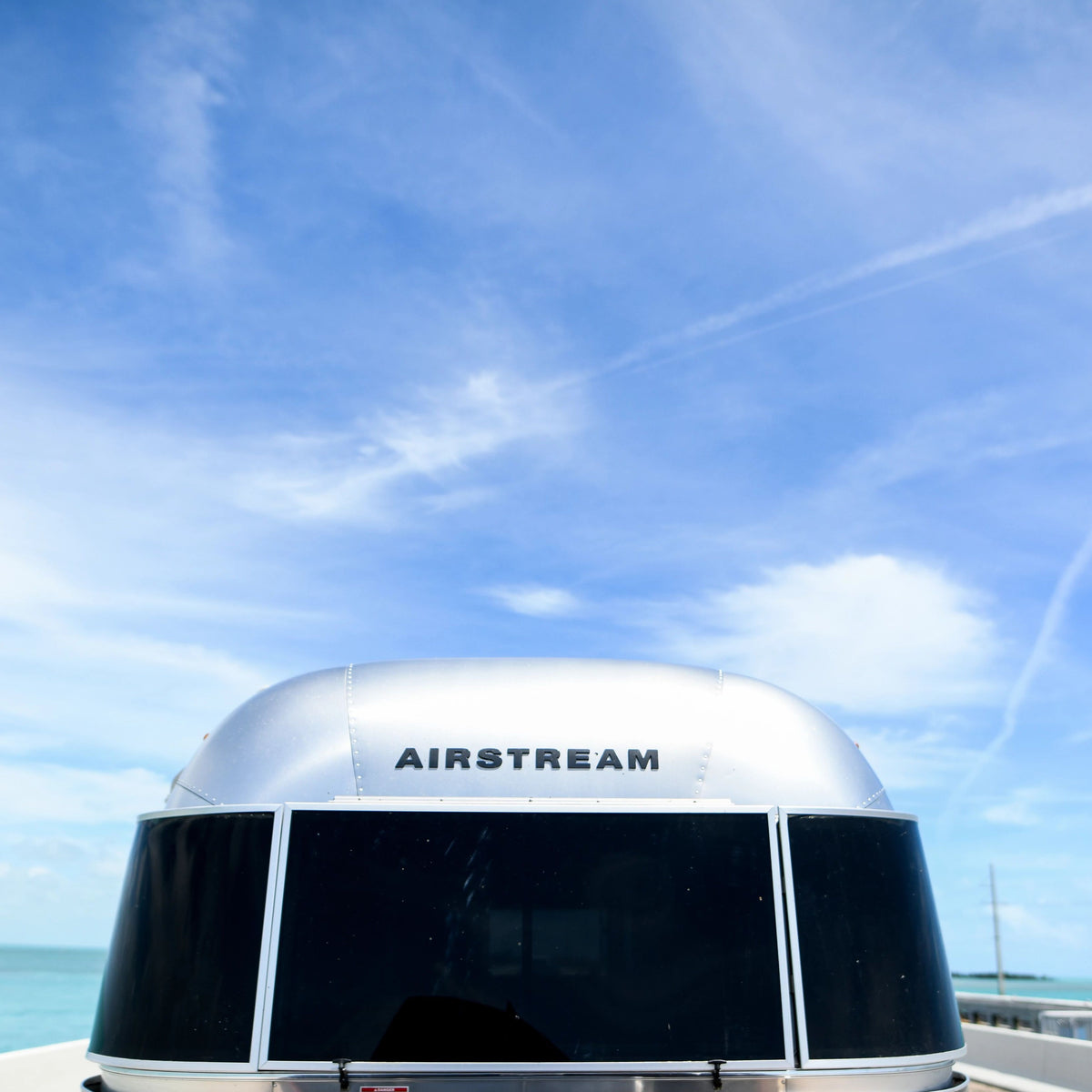 The front of an Airstream trailer with tinted windows is shown under a bright blue sky with wispy clouds, featuring the crisp Airstream Logo Decal in Black Chrome (Part #386272, 386205-01) that highlights the iconic name.