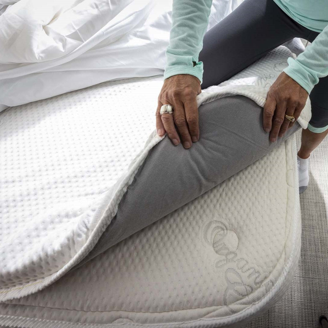 A person in a light blue shirt lifts the corner of an Airstream Memory Foam Topper for Safari Travel Trailers, revealing a gray mattress underneath. The bed is partially made with white sheets, creating a cozy setup perfect for an Airstream Safari Travel Trailer.