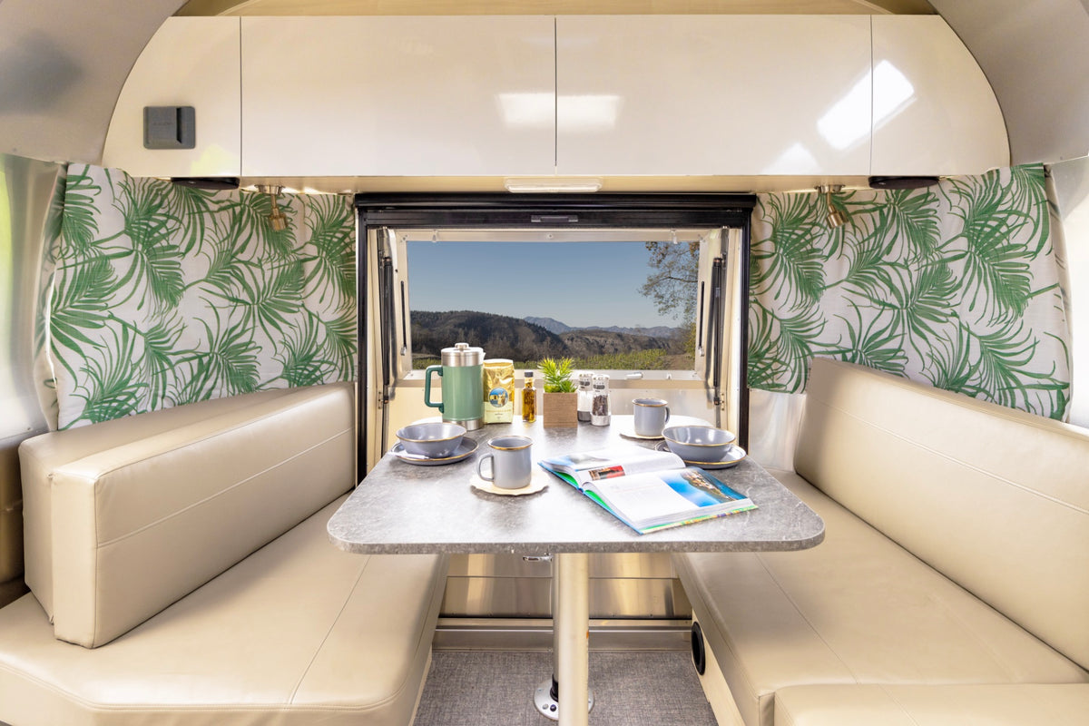 A cozy RV dining area with cream benches and a table set for four features Airstream Printed Custom Curtains for Flying Cloud Travel Trailers in a green palm leaf pattern, framing an open window that reveals a scenic mountain view beneath clear skies—perfectly complementing the inviting interior of this Airstream Flying Cloud.