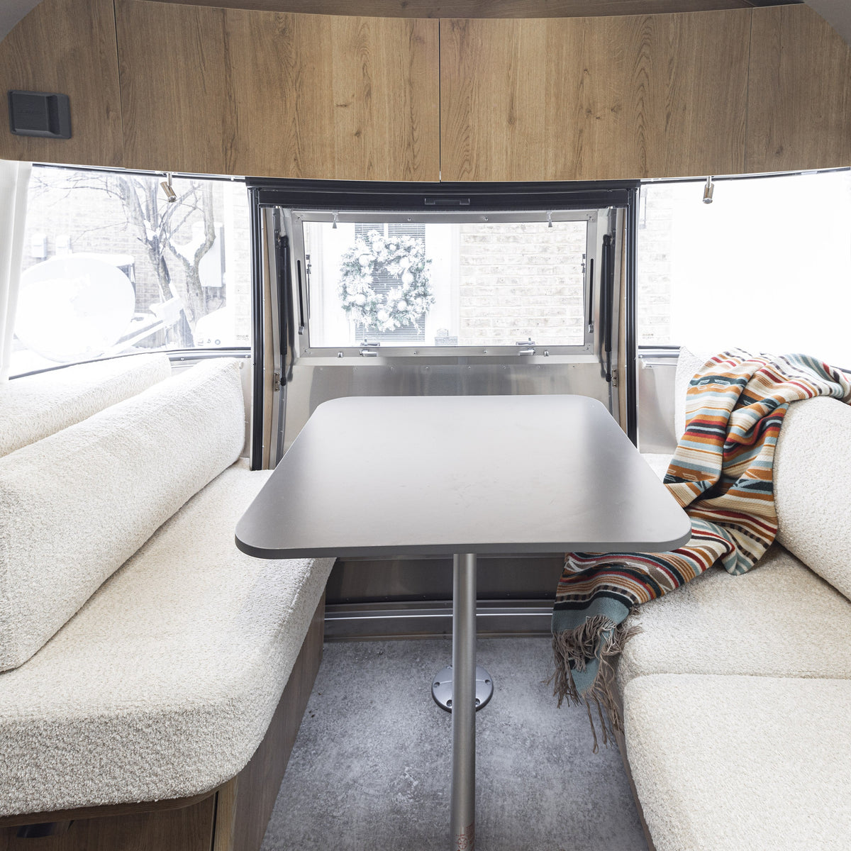 Interior of a camper with a table between two cushioned benches featuring Airstream Custom Replacement Cushions for 23′ CB International Travel Trailers for enhanced comfort, a colorful striped blanket draped on the right bench, and an open window at the end displaying a snowy outdoor scene.