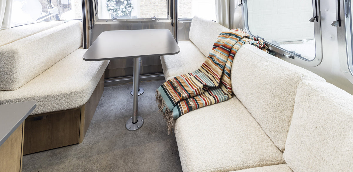 Modern RV interior with beige cushioned benches, a gray table, and a colorful striped blanket draped over one bench. Large windows provide natural light.