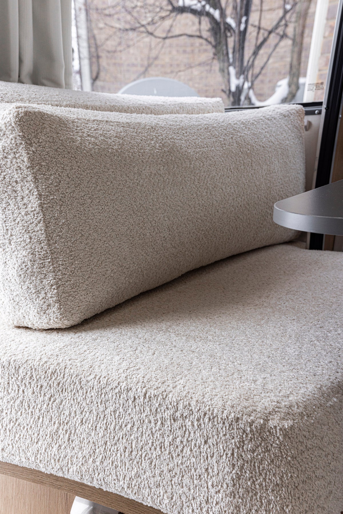 A close-up of a textured beige bench cushion with a matching rectangular pillow, set near a window framing a snowy tree and brick wall—perfect inspiration for Airstream Custom Replacement Cushions for 25′ and 27′ Flying Cloud Travel Trailers, blending both style and comfort in your space.