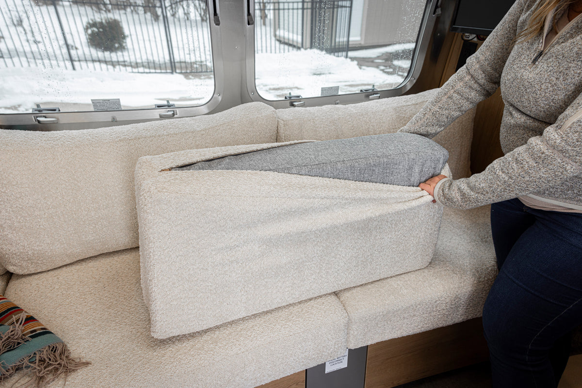 In an RV, someone unzips a beige cushion to reveal a gray inner layer, possibly examining the Airstream Custom Replacement Cushion Covers for Trade Wind Travel Trailers; snow can be seen outside the window in the background.