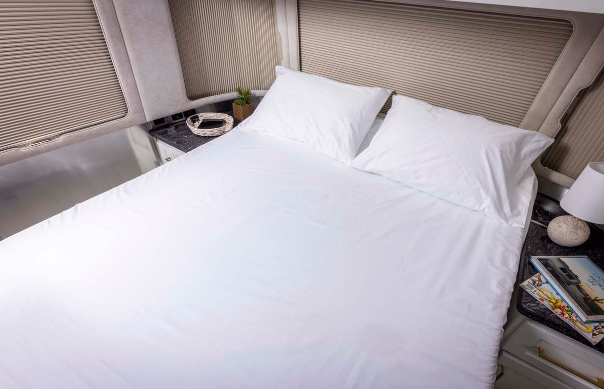 A neatly made bed with white sheets and pillows, next to nightstands holding a lamp, a basket, a small plant, and magazines, with window blinds behind the bed.
