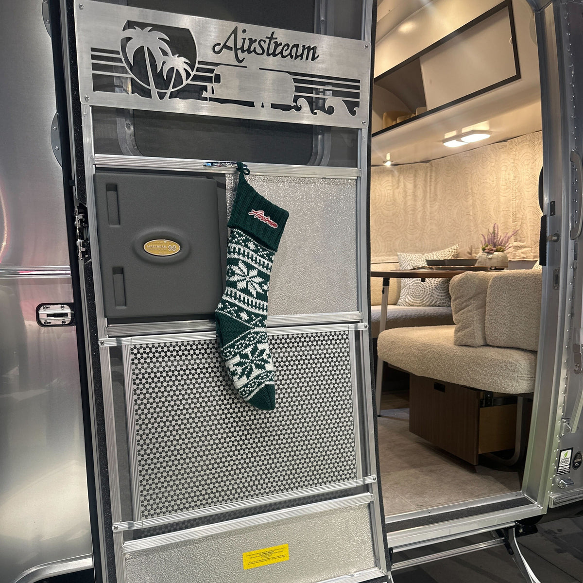 An Airstream Holiday Stocking in green and white hangs on the door of an Airstream trailer, bringing festive cheer to the scene. Inside, a cozy beige curved sofa and a small table with holiday décor are visible through the doorway.