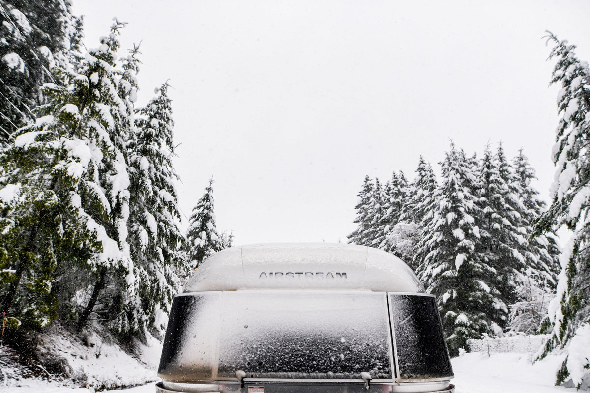 A snow-covered Airstream trailer is parked on a wintry road lined with tall evergreen trees blanketed in fresh snow.
