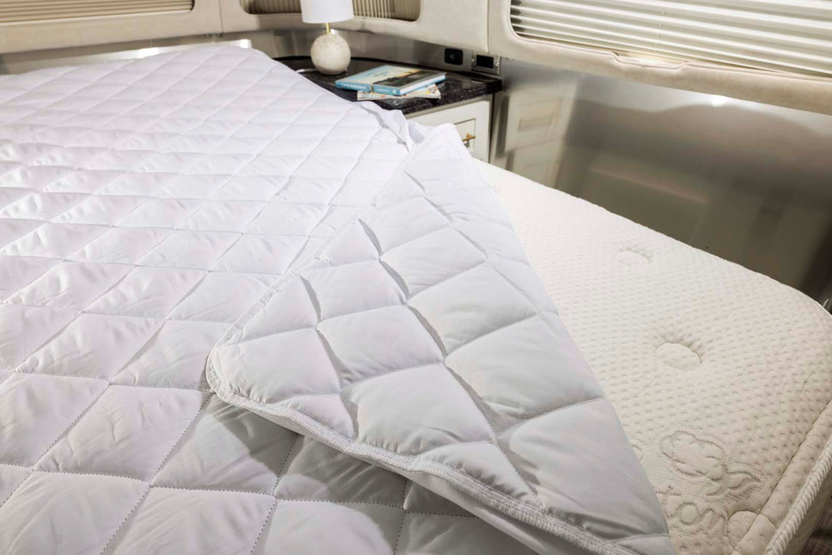 A close-up of a bed with a quilted white mattress protector partially pulled back, revealing a textured mattress underneath. There is a nightstand with books and a lamp in the background.