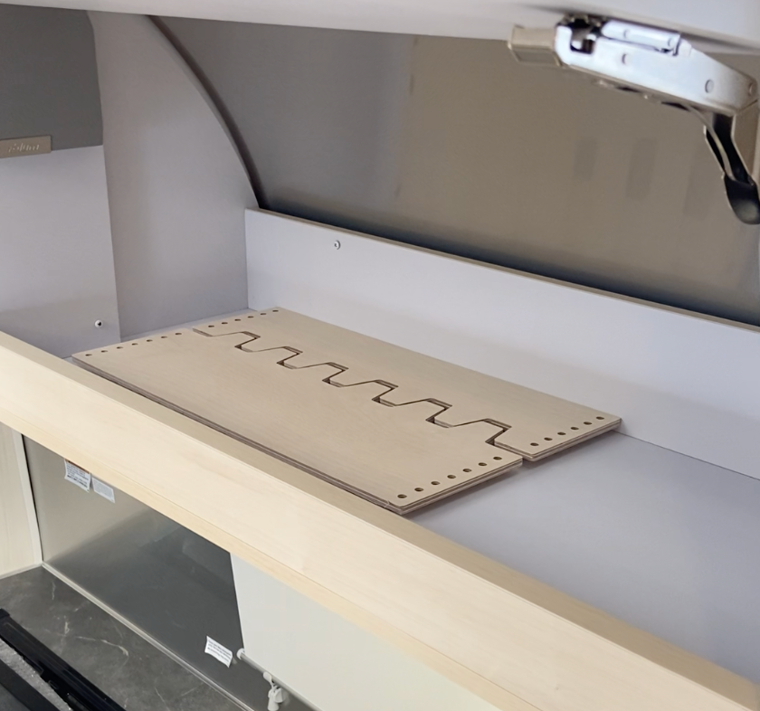 A flat wooden board with a wavy groove and small holes along the edges rests inside a clean, empty fume hood. The fume hood door is partially open, revealing the interior space and some visible labels.