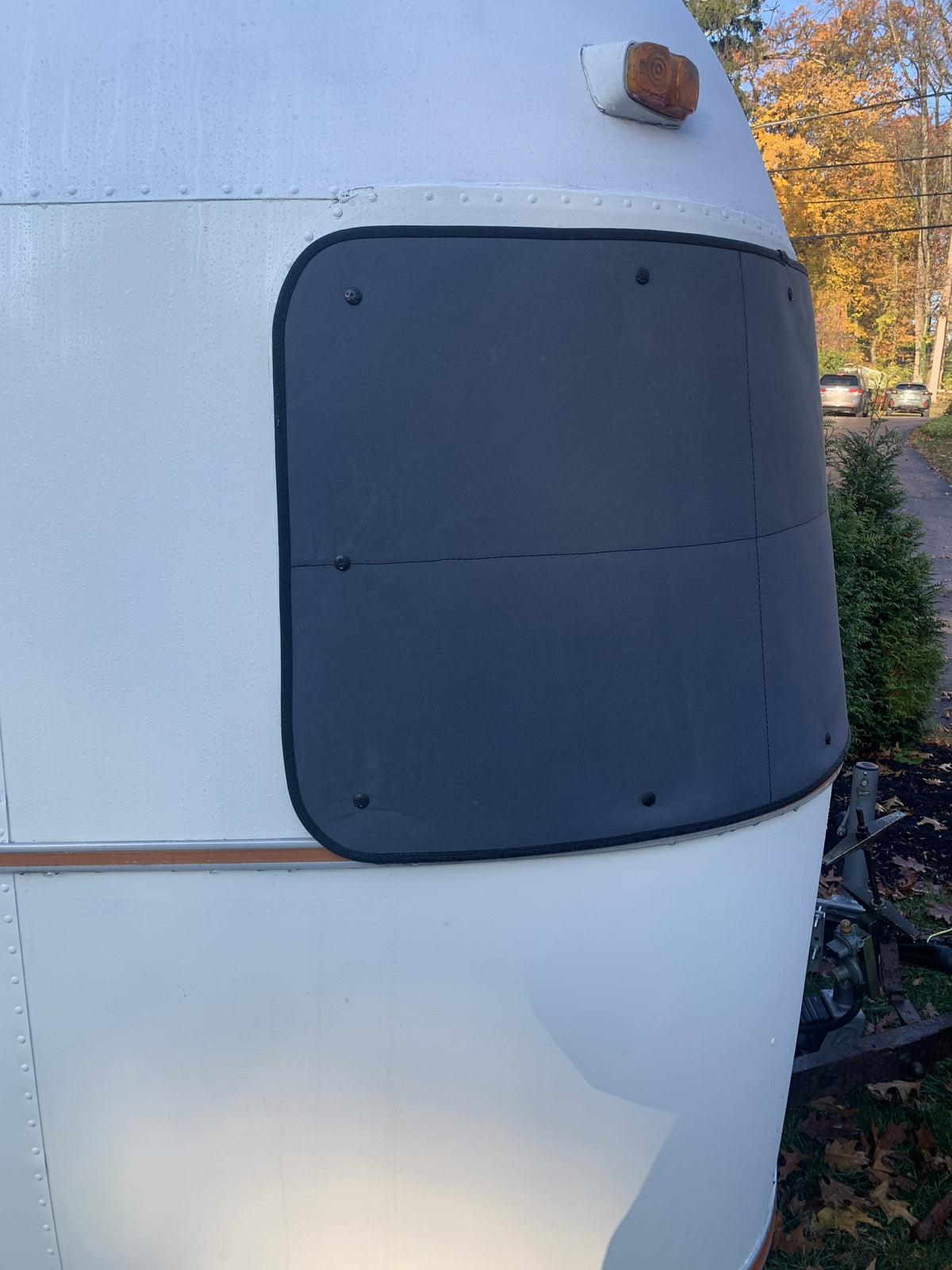 Close-up of a white trailer featuring the Airstream Padded Window Protector for Argosy—a large, black, rectangular cover over the window. An orange light is mounted above the window, and trees along with parked cars can be seen in the sunny background.