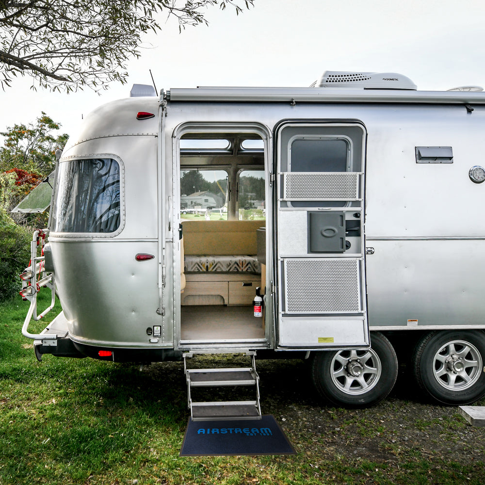 A silver Airstream trailer with an open door and steps leading inside, parked on grass. The interior shows a cushioned seating area. A mat reading AIRSTREAM is at the base of the steps.