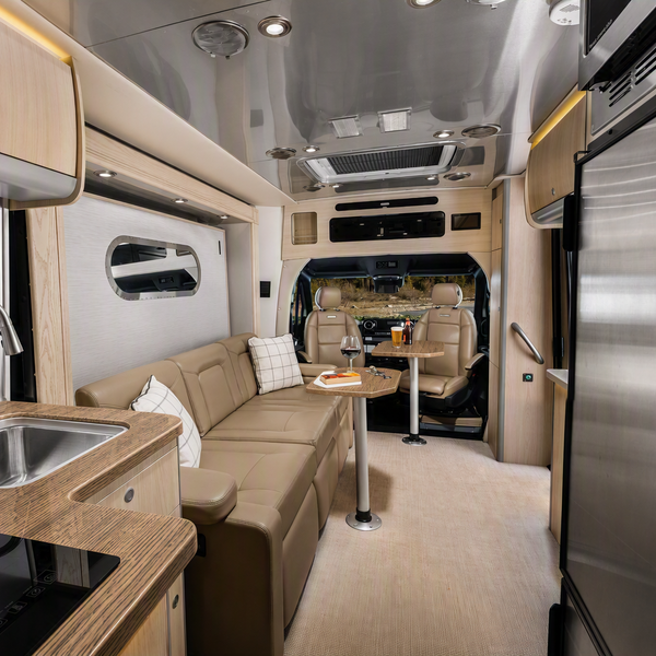 The interior of a recreational vehicle or camper, showing a kitchenette with a sink and cooktop, a beige sectional sofa, a small dining table, and stainless steel appliances including a refrigerator.