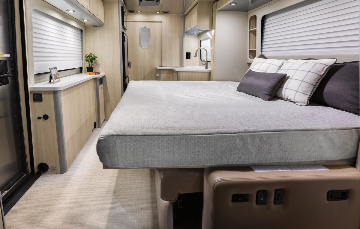Interior of a modern Airstream RV featuring a neatly made bed with the Airstream Fitted Sheet for Atlas 25MS Murphy Bed Cushion and plush pillows, surrounded by light wood cabinetry. The space also includes a small plant, ample countertop area, and a kitchen sink in the background. The décor is minimal and clean.
