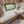 Compact recreational vehicle bedroom interior with a single bed, adjustable wooden table, storage drawers, shelving, and a window looking out onto grass.