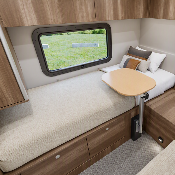 Compact recreational vehicle bedroom interior with a single bed, adjustable wooden table, storage drawers, shelving, and a window looking out onto grass.