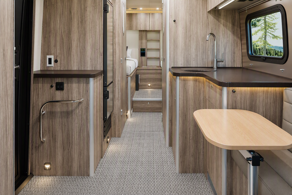 The interior of a recreational vehicle or camper, showing a compact kitchenette with wood grain cabinets, a sink, a small table in the foreground, and a carpeted aisle leading to a bathroom area.