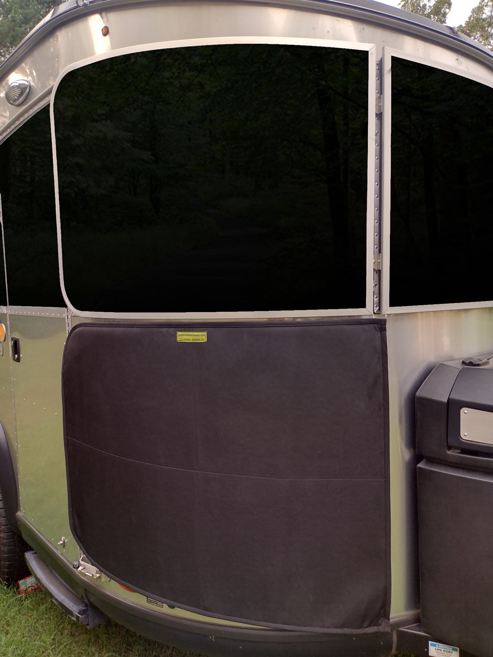 Close-up of a shiny silver Basecamp X Travel Trailer door featuring a large dark window and an Airstream Padded Stone Guard Cover for Basecamp X attached to the lower half for added rock guard protection, parked on green grass with trees reflected in the glass.