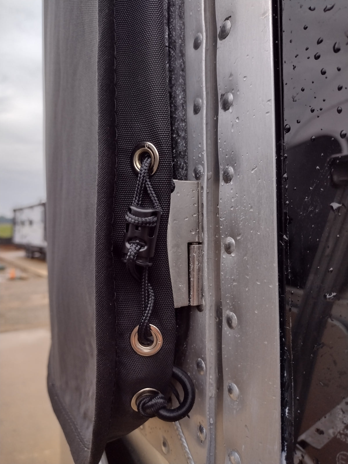 Close-up of the Airstream Padded Window Protector for Basecamp—a black tarp with metal grommets and a bungee cord, securely attached to the edge of a metal surface, likely an Airstream Basecamp travel trailer. Raindrops can be seen on the metal, highlighting how this window protector effectively shields your trailer’s windows during inclement weather.