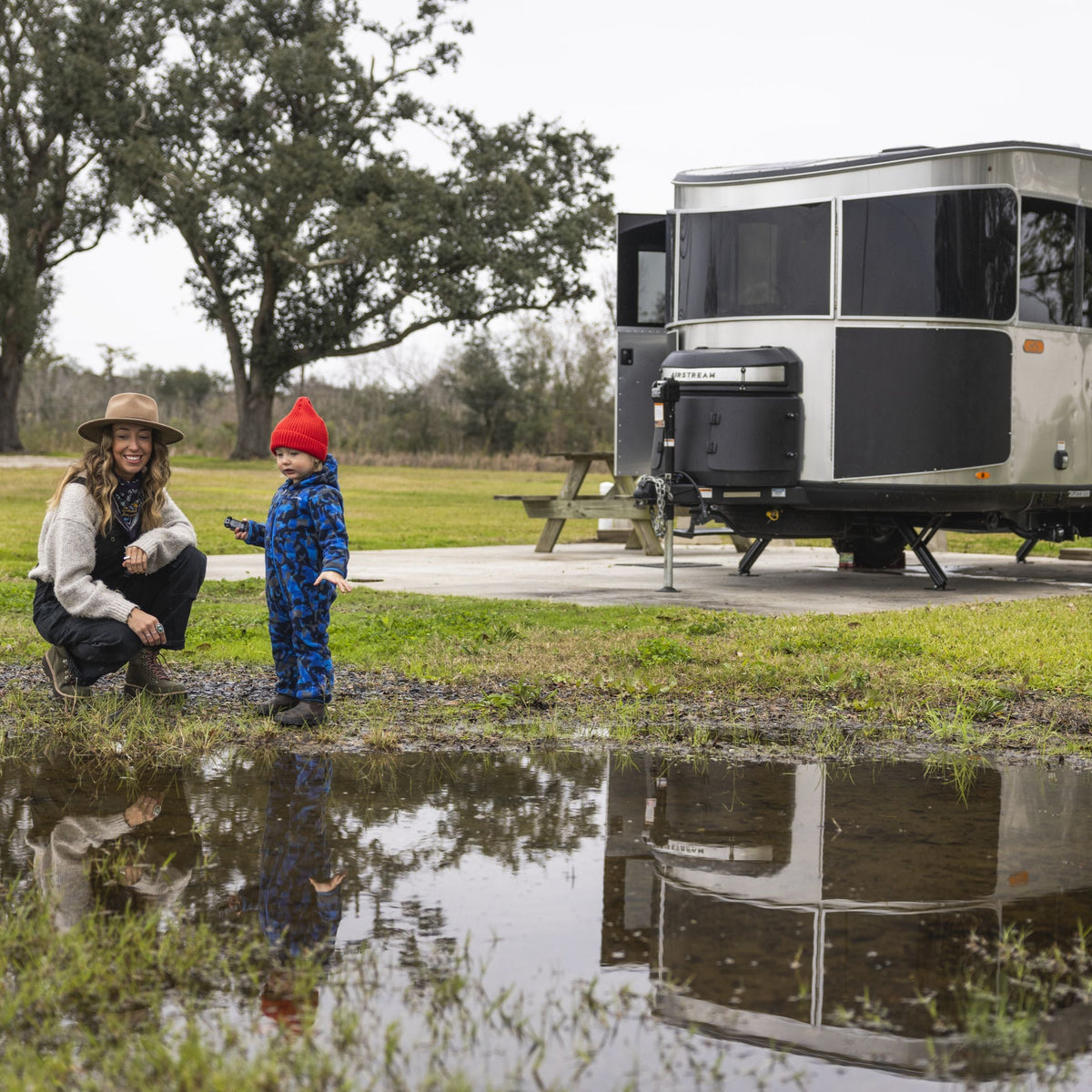 A woman and a child in blue overalls and a red hat stand by a puddle, with an Airstream Owner Manuals: Basecamp Xe Travel Trailer and picnic table in the background on a grassy, overcast day.