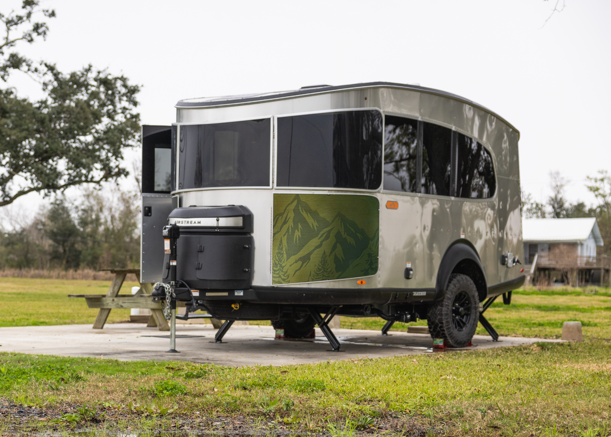A Basecamp X travel trailer featuring dark windows, a green mountain design, and the Airstream Rock Guard Rock Wraps Nature Collection for Basecamp X is parked on a concrete pad in a grassy field near trees and a wooden picnic table, with a small building in the background.